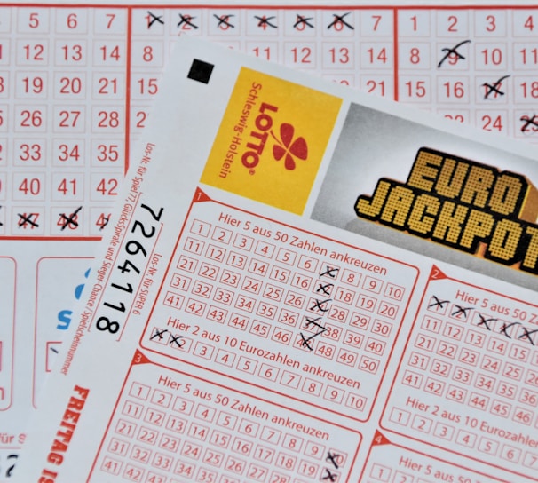 Three lottery tickets are displayed, including Eurojackpot and Super 6. Various numbers are marked on the tickets, with some crossed out and others checked. The Eurojackpot ticket is partially visible with the logo prominently displayed, and a ticket number is visible on one of the tickets.